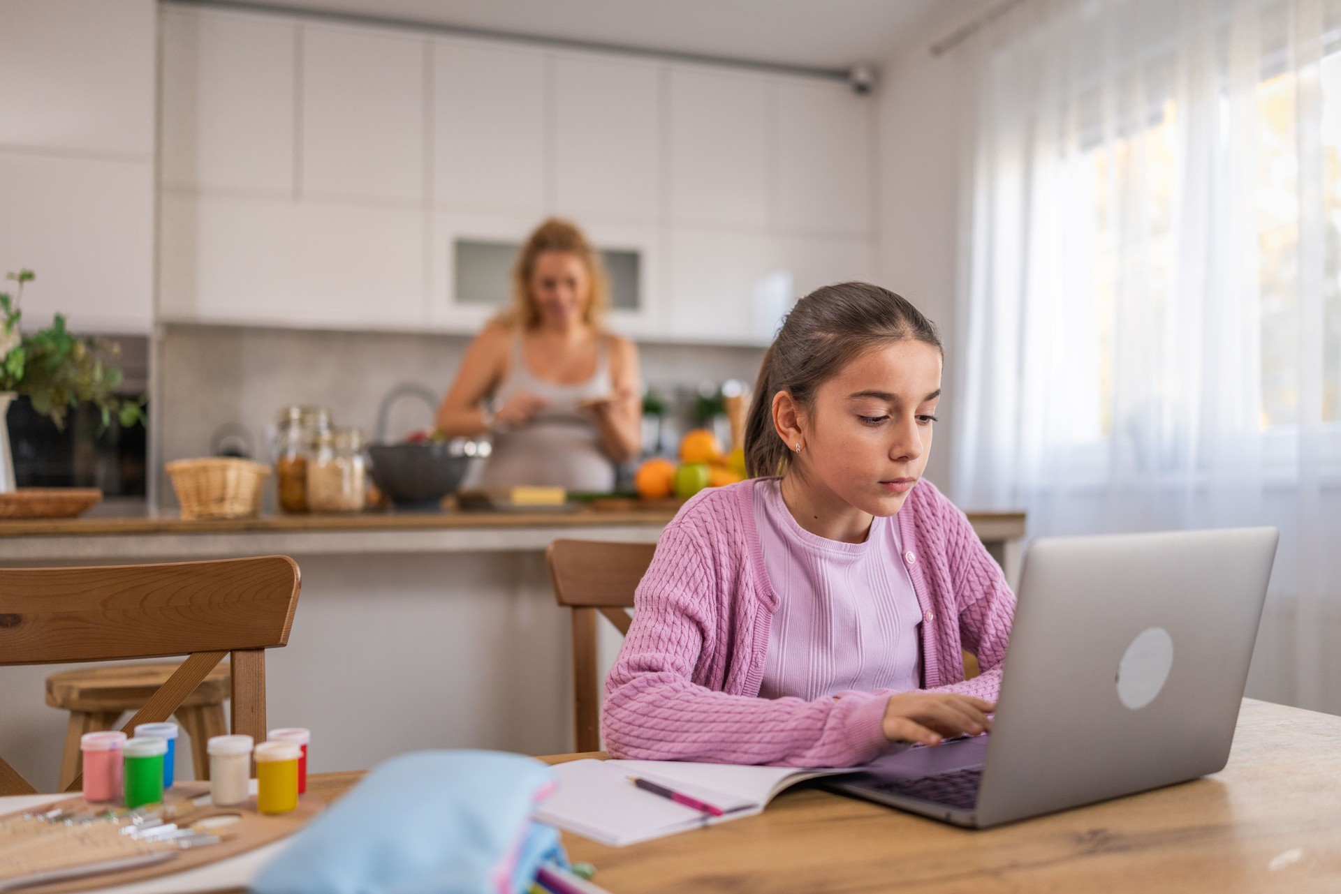 Cute girl sitting at dining table, doing homework online while her pregnant mother cooking breakfast in blurred background.